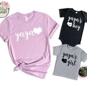 K&ouml;nnte beinhalten: Ein Set aus drei T-Shirts mit dem Text "yaya" und Herzen. Das T-Shirt f&uuml;r Erwachsene ist rosa, das T-Shirt f&uuml;r Jungen ist schwarz und das T-Shirt f&uuml;r M&auml;dchen ist grau.