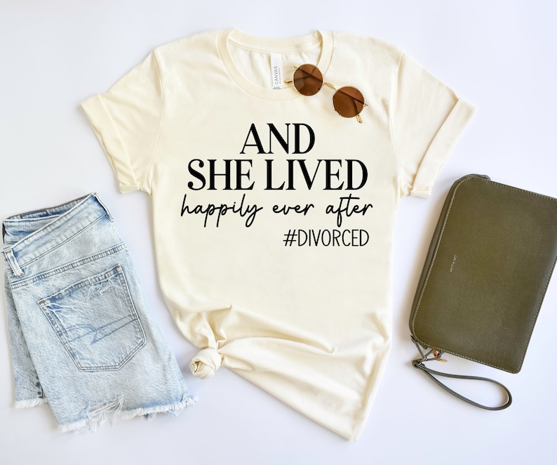 Puede incluir: Una camiseta blanca con el texto "AND SHE LIVED happily ever after #DIVORCED" impreso en negro.