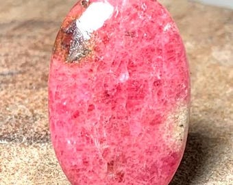 May include: A pink and white oval cabochon gemstone with a rough, textured surface. The stone has a pink hue with white inclusions.