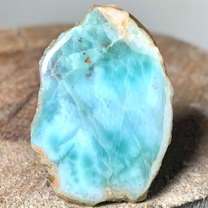 May include: A polished, irregular-shaped gemstone with a striking blue and turquoise color palette. The stone has a marbled appearance with areas of lighter and darker shades, and a rough, natural edge. Small flecks of brown are visible.