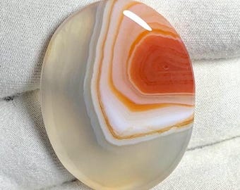 Pode incluir: Gema ovalada com superfície polida. A pedra apresenta faixas concêntricas de cor, incluindo tons de laranja, branco e cinza. O centro da pedra tem um tom laranja vibrante, criando um contraste visual marcante. A gema é provavelmente usada para joias ou fins decorativos.