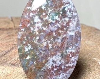 May include: Oval-shaped gemstone with a polished surface. The stone features a mix of colors, including red, white, green, and brown, creating a unique pattern. The gemstone is set against a wooden background, highlighting its natural beauty.