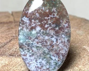 May include: An oval-shaped gemstone with a polished surface. The stone features a mix of colors, including red, green, and white, with a speckled pattern. The gemstone is set against a wooden surface.