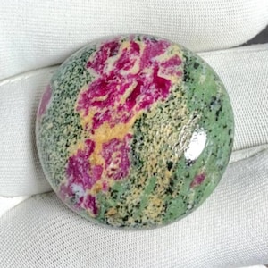 May include: A round gemstone with a polished surface. The stone features a vibrant pattern of magenta and yellow hues set against a speckled green background. Small black flecks are scattered throughout the stone, adding depth and detail.