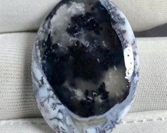May include: An oval-shaped gemstone with a unique pattern of black, gray, and white swirls. The polished surface reflects light, highlighting the natural variations within the stone. The gemstone's design resembles a landscape or cloud formation.