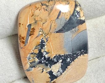 May include: A polished, light brown, gray, and black jasper cabochon with a unique, swirling pattern. The stone has a smooth, polished surface and is a beautiful example of this type of gemstone.
