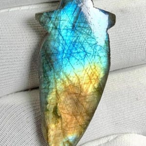Puede incluir: Una piedra preciosa de labradorita pulida, con forma de punta de flecha con una parte superior en forma de estrella. La piedra muestra un vibrante juego de colores, con azules, verdes y amarillos. La superficie tiene una red de líneas finas.