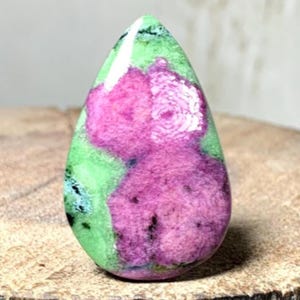 Peut inclure: Une pierre précieuse polie en forme de goutte d'eau, présentant un mélange de couleurs vives. La pierre présente une zone proéminente rose foncé, entourée d'une teinte verte claire. De petits points noirs sont dispersés dans la pierre.