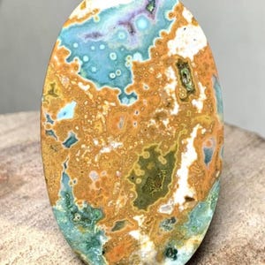 May include: Oval-shaped gemstone with a unique pattern of colors. The stone features a blend of blue, orange, green, and white hues, creating a marbled effect. The polished surface highlights the natural variations and textures of the stone.
