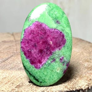 Może przedstawiać: Owalny kamień szlachetny z żywą zieloną podstawą i wyraźnym, purpurowym środkiem. Wypolerowana powierzchnia kamienia ukazuje naturalne wzory. Kamień szlachetny jest prezentowany na drewnianej powierzchni, podkreślając jego kolory i tekstury.