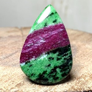 Op de afbeelding: Een gepolijste, druppelvormige edelsteen met een opvallende kleurencombinatie. De steen heeft een levendige groene basis met donkergroene spikkels, doorsneden door een band van diep bordeauxrood en wit. De edelsteen ligt op een houten oppervlak.