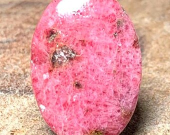 May include: A pink and white cabochon gemstone with a rough, textured surface. The stone has a pink and white color with a hint of gold.