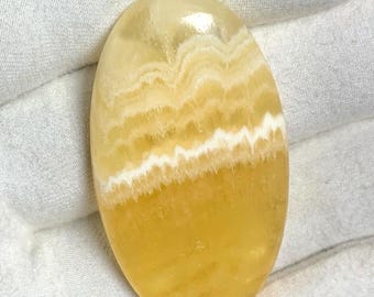 Puede incluir: Una piedra preciosa ovalada en cabujón con bandas amarillas y blancas. La piedra tiene una superficie lisa y pulida y está colocada sobre un fondo blanco.