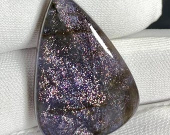 May include: A polished, teardrop-shaped gemstone with a dark purple base and shimmering, glitter-like inclusions. The stone has a smooth, glossy surface. The gemstone's unique pattern and colour make it a striking piece, set against a white background.