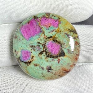 May include: A round gemstone with a mottled surface. The stone features vibrant pink inclusions set against a background of green, brown, and yellow hues. The polished surface reflects light, highlighting the stone's natural patterns and colors.