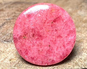 May include: A round, pink, polished gemstone with a speckled texture. The stone is approximately 1 inch in diameter.