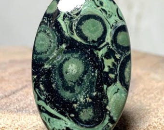 May include: Oval-shaped gemstone with a striking pattern of green and black swirls and circular designs. The polished surface reflects light, highlighting the natural variations in color and texture. The stone is displayed on a wooden surface.