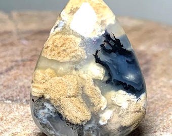 May include: A teardrop-shaped gemstone with a polished surface. The stone features a blend of colors, including beige, black, white, and blue, creating a unique, natural pattern. The stone is likely used for jewelry or display.