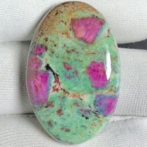 Könnte beinhalten: Ovaler Edelstein mit polierter Oberfläche. Der Stein weist eine Mischung aus Farben auf, darunter leuchtendes Pink, helles Grün und Braun. Die rosa Bereiche haben ein kristallines Aussehen, während die grünen und braunen Bereiche eine gesprenkelte Textur aufweisen. Der Edelstein liegt vor weißem Hintergrund.