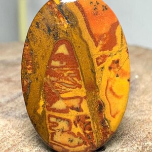 Pode incluir: Um cabochão de pedra preciosa polida de formato oval com uma mistura vibrante de cores quentes, incluindo amarelo mostarda, laranja queimado e castanho escuro. A pedra exibe padrões naturais intrincados e manchas escuras na superfície.