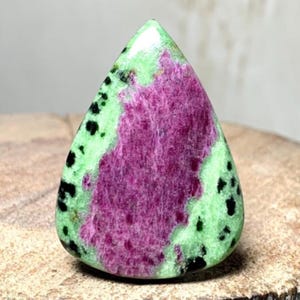 May include: A teardrop-shaped gemstone with a polished surface. The stone features a vibrant mix of colors, including a central area of deep magenta, surrounded by a light green hue speckled with black spots. The gemstone is displayed on a wooden surface.
