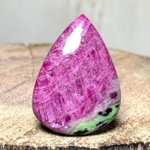 May include: A polished, teardrop-shaped gemstone with a vibrant pink hue, showcasing a textured surface. The stone features a band of green and black near the base, set against a natural wood grain background.