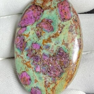 Puede incluir: Gema ovalada con superficie pulida. La piedra presenta una mezcla de colores, incluyendo rosa vibrante, verde claro y marrón. Las áreas rosas están agrupadas, creando un patrón único. La gema se sostiene sobre un fondo blanco.
