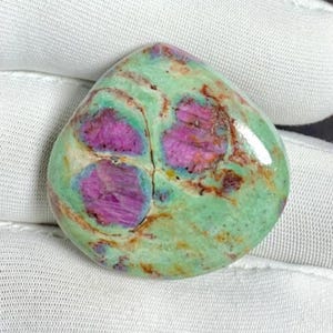 Puede incluir: Una piedra preciosa pulida con forma redondeada e irregular. La piedra presenta un color base verde claro con inclusiones de color magenta vibrante. Vetas marrones y tostadas recorren la piedra, lo que aumenta su apariencia natural.