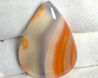 May include: A teardrop-shaped agate cabochon with a white, gray, and orange banded pattern. The stone has a smooth, polished surface.