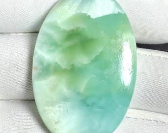Può includere: Pietra preziosa ovale con superficie lucida. La pietra presenta una miscela di tonalità verde chiaro e turchese, con un aspetto nuvoloso e marmorizzato. La gemma è posta su uno sfondo di tessuto bianco.