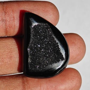 May include: Black drusy agate cabochon with a smooth, polished surface and a sparkly, crystalline interior. The cabochon is shaped like a teardrop and has a dark, almost black, color.