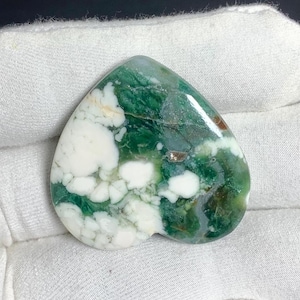 Può includere: Una pietra preziosa lucida a forma di goccia con un motivo marmorizzato di verde bosco intenso e sezioni bianco opaco. La superficie è liscia e riflettente, mostrando inclusioni naturali e varie transizioni di colore sulla faccia della pietra.