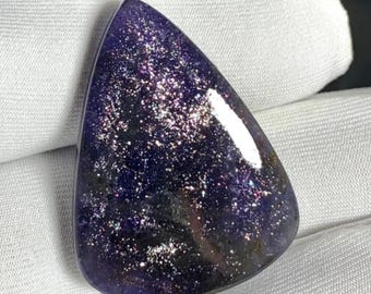 May include: A polished, teardrop-shaped gemstone with a deep purple hue, speckled with shimmering silver and iridescent flecks. The stone has a smooth, glossy surface and a dark base.