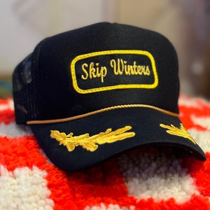 Könnte beinhalten: Schwarze Trucker-Kappe mit einem goldenen gestickten Aufnäher mit der Aufschrift "Skip Winters". Die Kappe hat auch goldene Stickdetails am Schirm.
