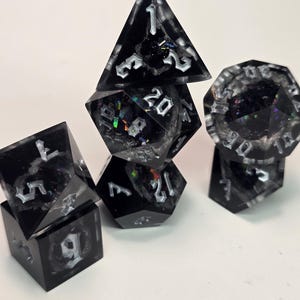 May include: A set of black resin dice with shimmering internal details and white numerals. The dice are in various shapes, including a pyramid, a cube, and a 20-sided die. The dice are stacked on a white surface, suitable for tabletop games.