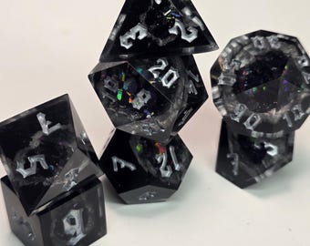 Könnte beinhalten: Eine Sammlung schwarzer Harzwürfel mit schillernden Einschlüssen und weißen Zahlen. Die Würfel haben verschiedene geometrische Formen, darunter eine Pyramide, ein Würfel und ein 20-seitiger Würfel. Sie sind auf einer weißen Oberfläche gestapelt.