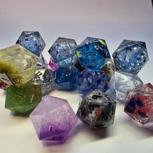 May include: A collection of polyhedral dice in various colors, including blue, green, purple, and clear. Some dice have glitter or embedded designs. The dice are made of a clear resin material and are used for tabletop games.