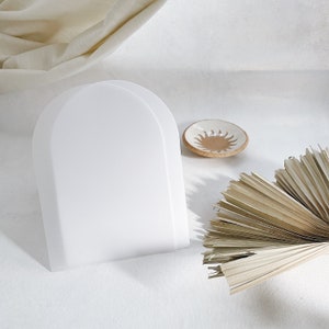 May include: A white, arched display stand dominates the image, alongside a small, round ceramic dish featuring a sunburst pattern. Dried palm leaves provide a natural contrast. The items are arranged on a soft, white surface, creating a minimalist aesthetic.