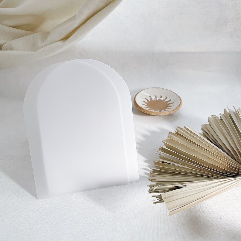 May include: A white, arched display stand dominates the image, alongside a small, round ceramic dish featuring a sunburst pattern. Dried palm leaves provide a natural contrast. The items are arranged on a soft, white surface, creating a minimalist aesthetic.