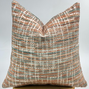 May include: Square cushion with a woven textured pattern. The cushion features a grid design in shades of orange, teal, and white. A home decor item.