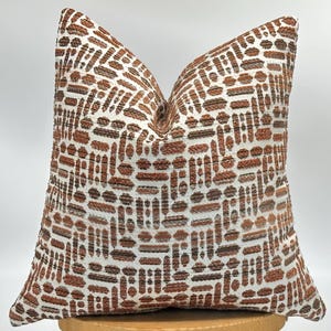 May include: A patterned cushion in brown and white. The design features a repeating geometric pattern of rectangles and ovals. The cushion is designed to add a decorative touch to any room. The cushion measures approximately 45cm x 45cm.