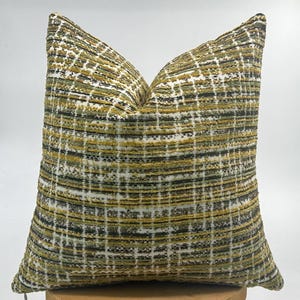 May include: Square cushion with a textured, woven fabric. The cushion features a grid pattern in shades of olive green, black, and white. The cushion is placed on a wooden surface.