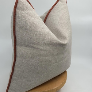 Puede incluir: Una almohada de tiro gris claro con un borde de color óxido. La almohada está sentada en un soporte de madera.