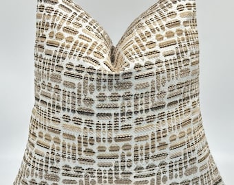 May include: A decorative square pillow with a textured pattern in shades of beige, brown, and white. The pillow has a woven design with a geometric pattern, and a zipper closure. The pillow is placed on a wooden surface.