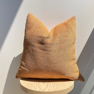 May include: A brown throw pillow with a textured fabric. The pillow is sitting on a wooden stool.