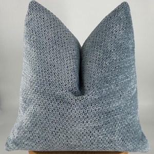 May include: A large, square, light blue decorative pillow. The pillow has a textured, woven appearance and is made of a soft, plush material. The pillow is resting on a wooden surface and is against a white background.