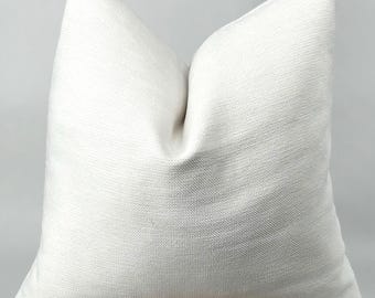 Op de afbeelding: Een vierkant sierkussen in een effen gebroken witte kleur met een duidelijk zichtbare, geweven textuur. Het kussen heeft een gestructureerde vorm met puntige hoeken en een licht ingedeukt midden, wat zorgt voor een strak en eenvoudig ontwerp.