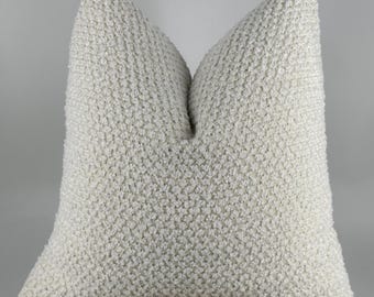 May include: A textured, off-white cushion with a soft, nubby surface. The cushion is square-shaped and appears to be made of a woven fabric. It is placed on a wooden surface.