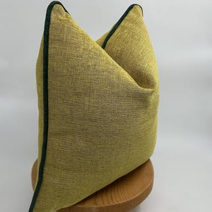May include: A yellow throw pillow with a dark green trim. The pillow is sitting on a wooden stand.