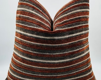 May include: A square decorative pillow with a striped pattern. The stripes alternate between shades of brown, gray, and off-white. The pillow has a soft, textured appearance, suggesting a plush fabric. The pillow is likely for home decor.
